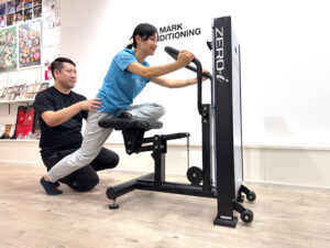 ZERO-iマシンで前傾姿勢をとる女性を、トレーナーが腰を支えながら可動域を広げるストレッチをしている様子。
