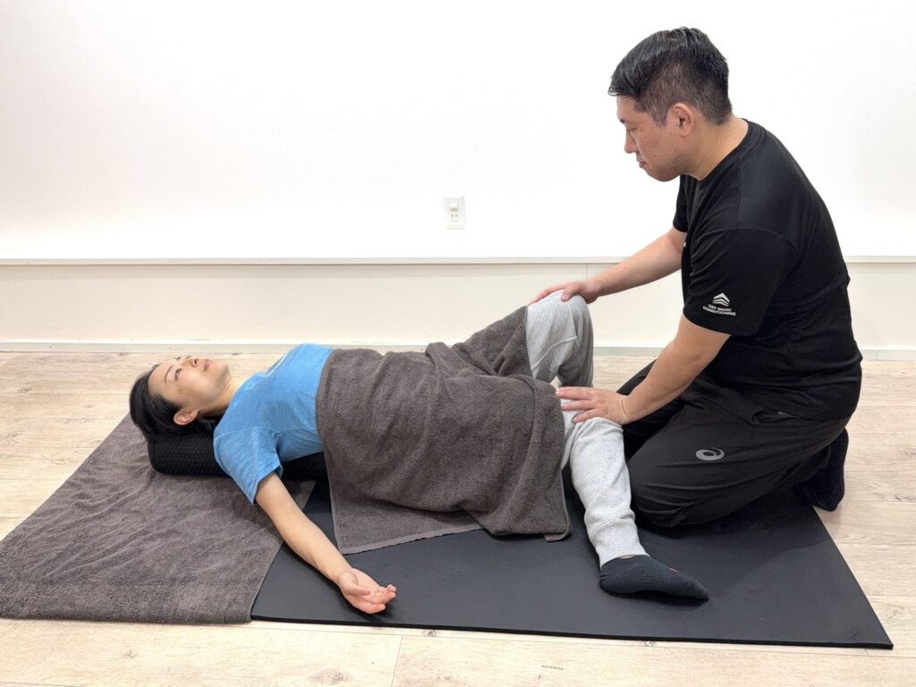女性が仰向けで膝を曲げ、トレーナーが股関節まわりをサポートしてストレッチしている様子。