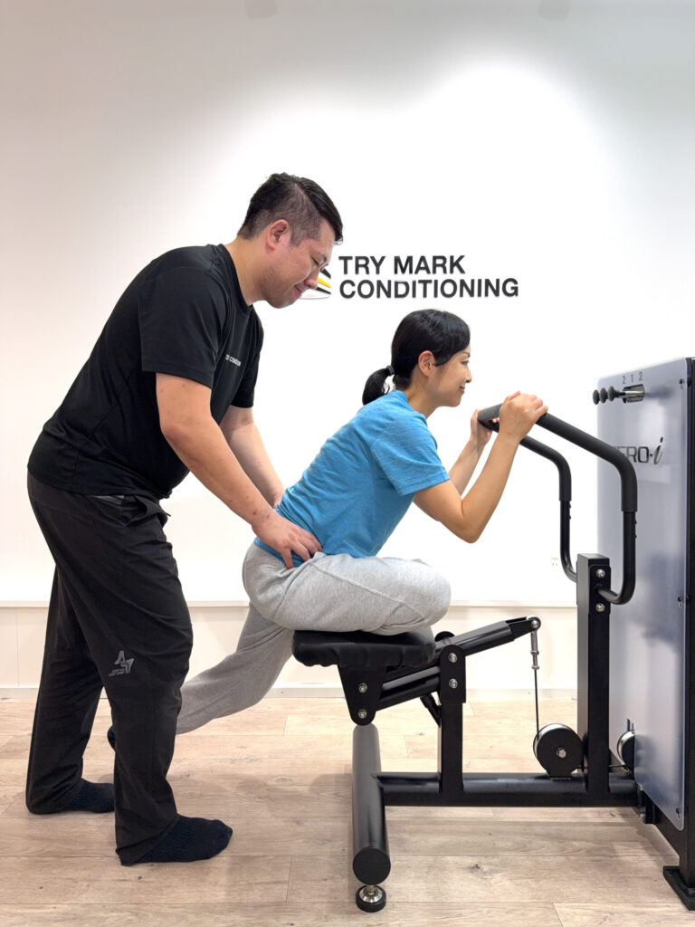 女性がZERO-iマシンで体を前傾させ、トレーナーが腰を支えながら動作をサポートしている様子。