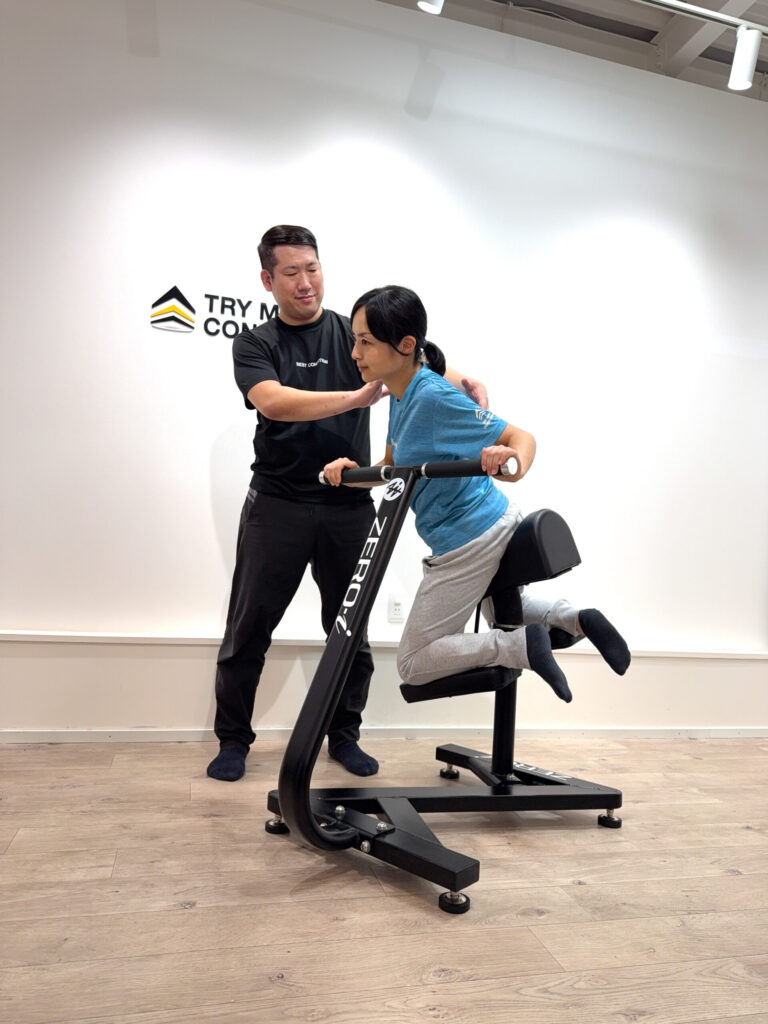 女性がZERO-i体幹マシンで前傾姿勢を保ち、トレーナーが肩を支えながら安定をサポートしている様子。