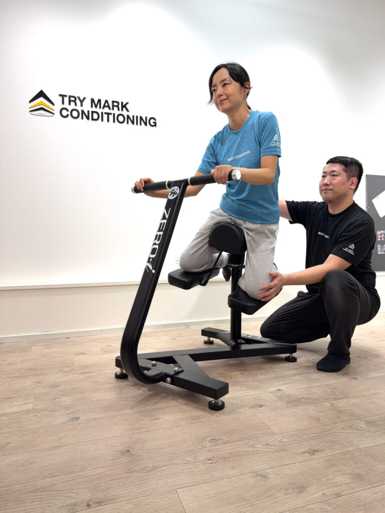 ZERO-iマシンで体幹トレーニングを行う女性。トレーナーが横から腰を支えて動作を補助している様子。