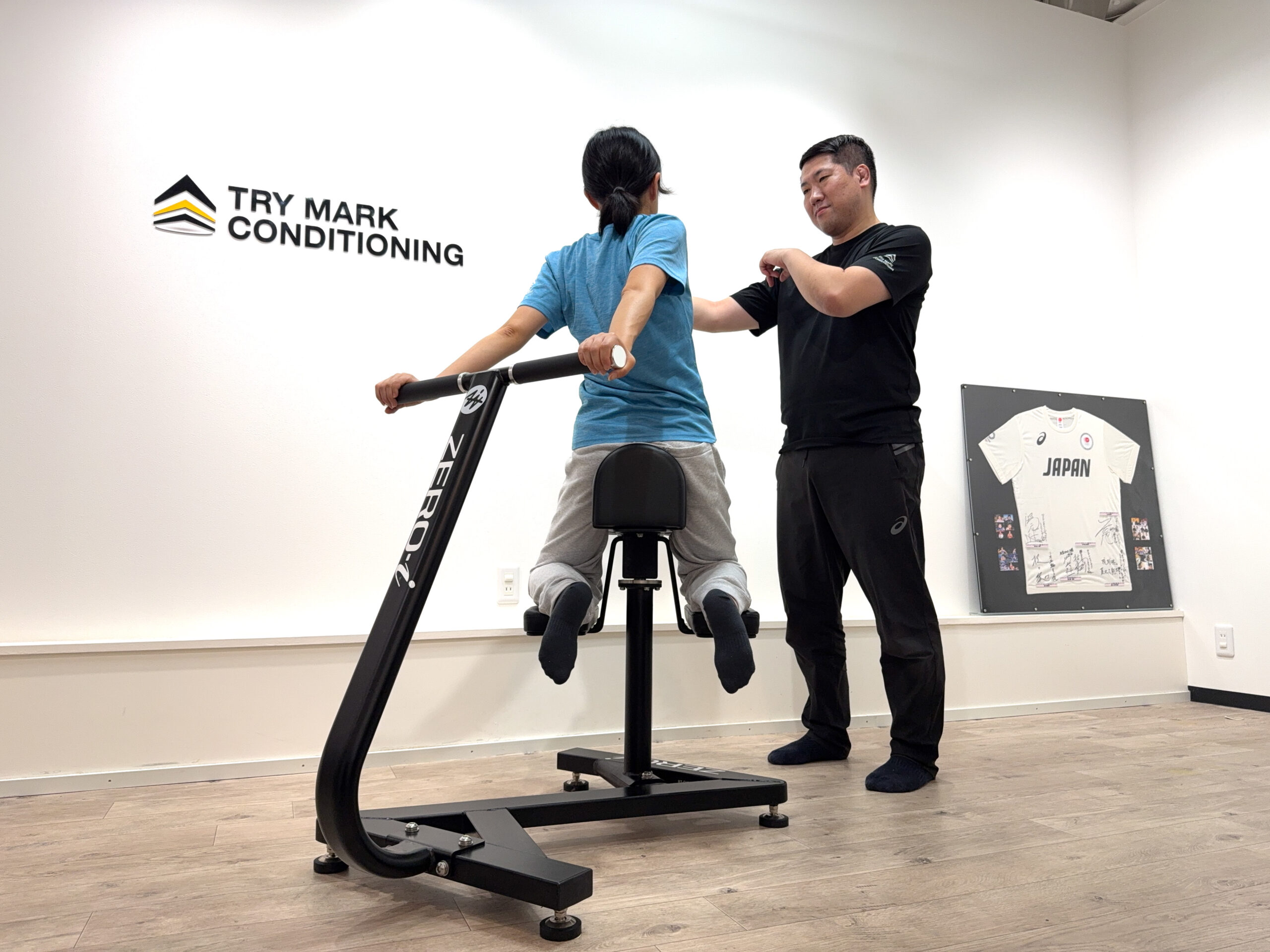 ZERO-iマシンで女性が背中を反らせ、トレーナーが肩に手を添えて正しい姿勢を指導している様子。
