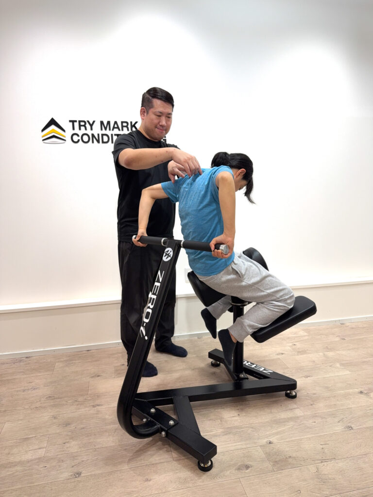 ZERO-iマシンで女性が胸を張って後方に反る動作を行い、トレーナーが肩の位置を調整している様子。