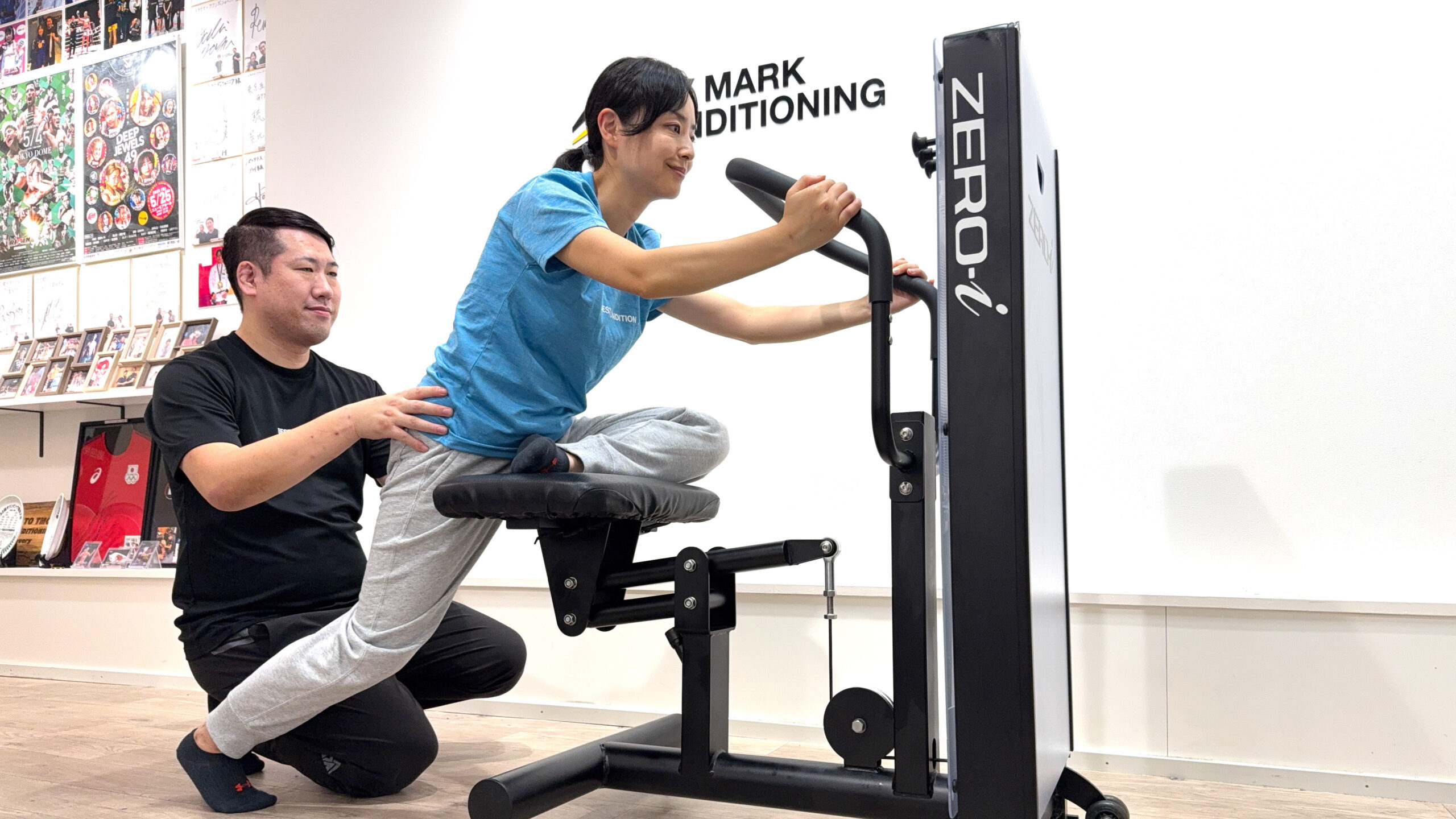 ZERO-iマシンで前傾姿勢をとる女性を、トレーナーが腰を支えながら可動域を広げるストレッチをしている様子。